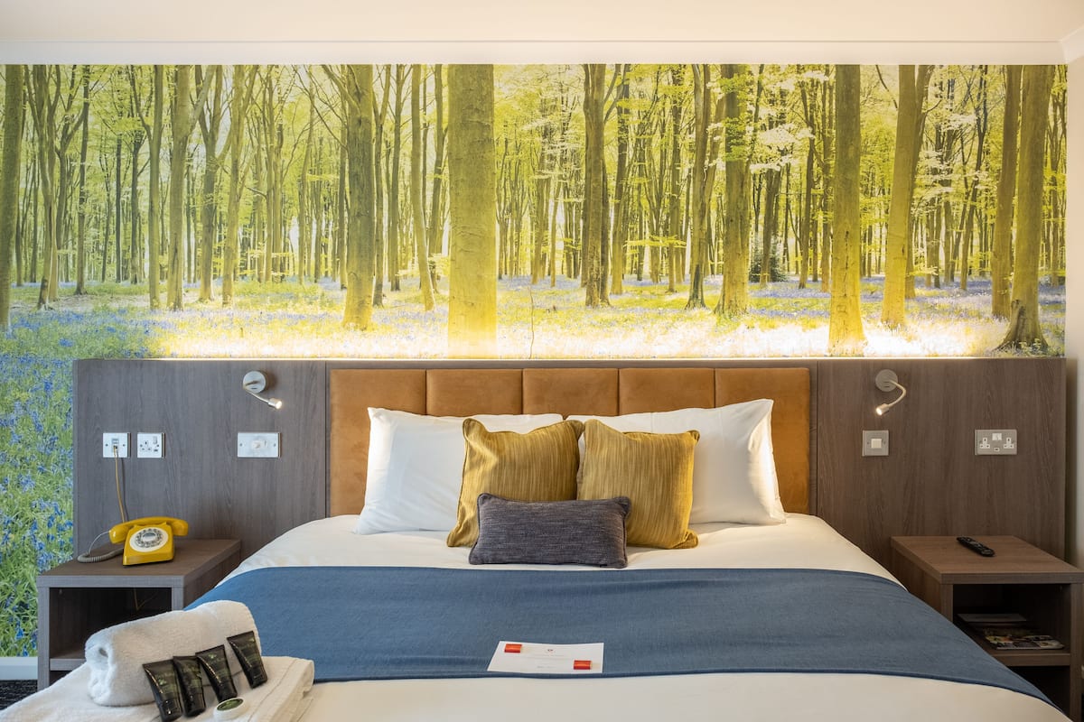 Camera d'albergo con un letto grande, una testiera, un comodino, un telefono, un telecomando, un asciugamano e un biglietto di benvenuto.