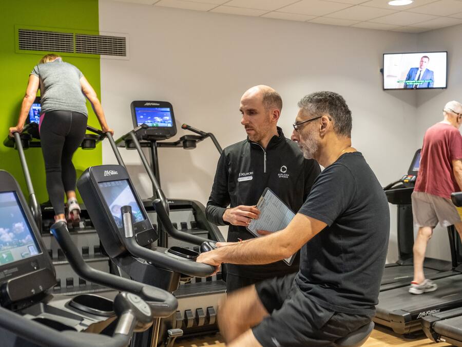 A gym with treadmills, a woman using one, and a man holding a clipboard.
