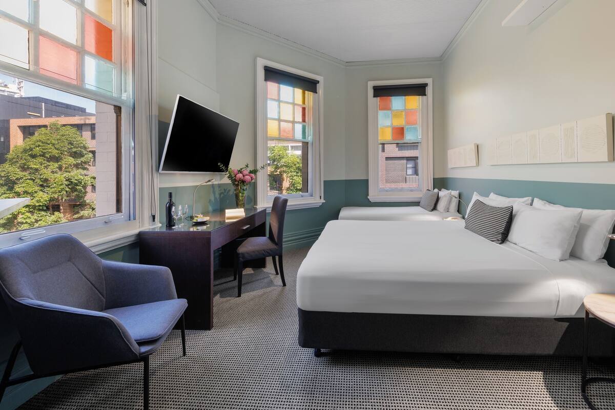 A hotel room with a bed, a desk, a chair, a TV, and large windows with colorful views.