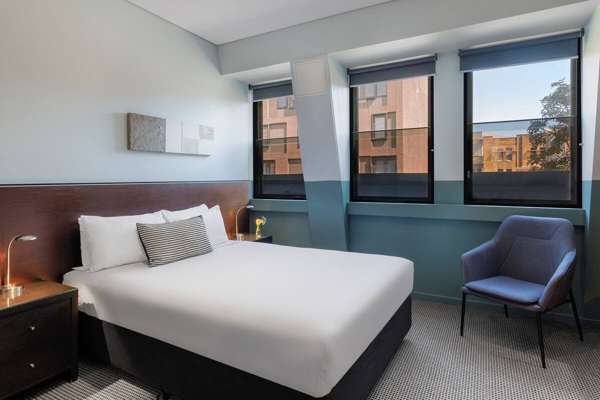 A hotel room with a large bed, a chair, a nightstand with a lamp, and a window with a view of buildings.