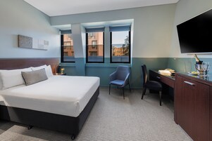 Habitación de hotel con cama, escritorio, silla, televisor y ventana con vista a edificios.