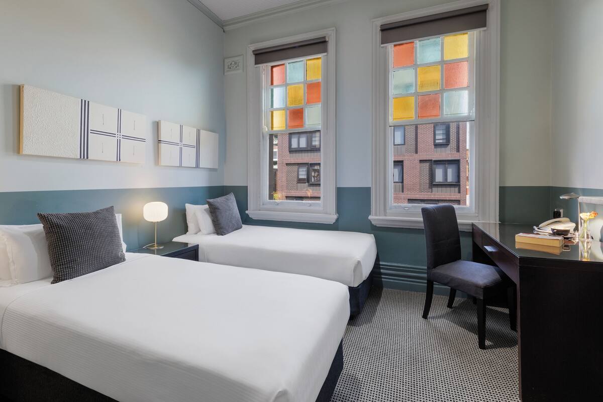 A hotel room with two beds, a desk, a chair, a lamp, and a view of buildings through windows.