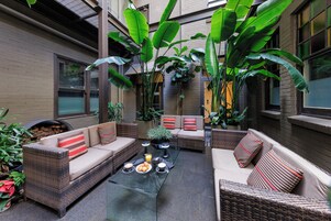 Un patio con mobiliario de mimbre, una mesa de vidrio y plantas verdes grandes.