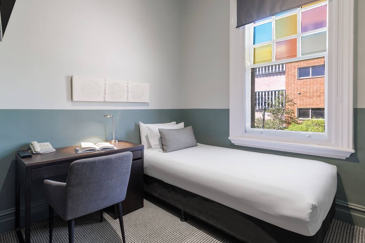 A bedroom with a bed, a desk, a chair, and a window with a view of a brick building.