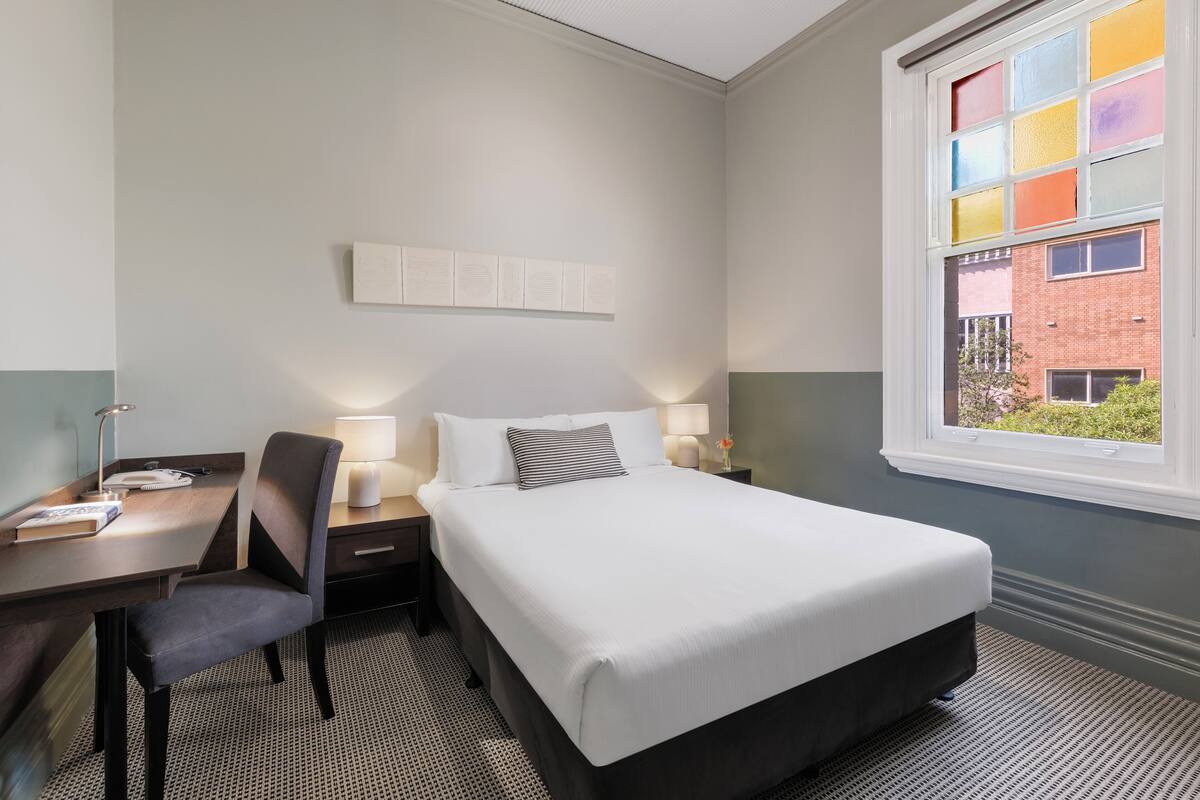 A hotel room with a bed, desk, chair, and a window with a view of a building.