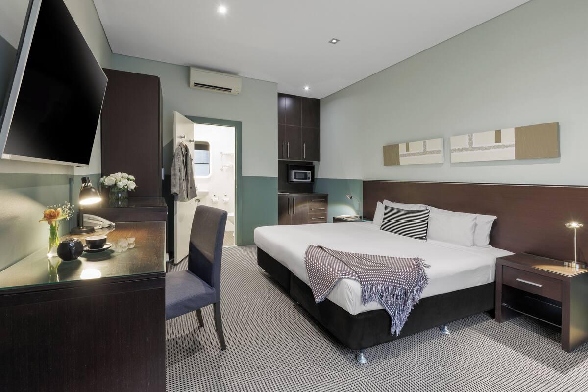 A hotel room with a bed, bedside tables, a desk with a chair, a TV, a microwave, and a wardrobe.