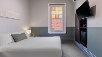 A hotel room with a bed, a TV mounted on the wall, a window with a view of a brick wall, and a small table with a lamp.