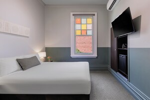 Et hotelværelse med en seng, et fjernsyn ophængt på væggen, et vindue med udsigt til en murstensmur samt et lille bord med en lampe.