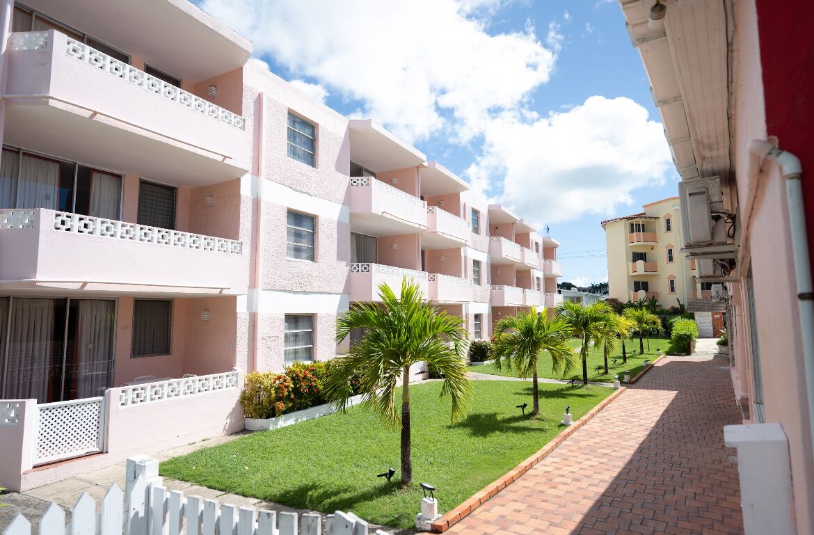 A multi-story hotel with balconies, a paved walkway, and a well-maintained lawn with palm trees.