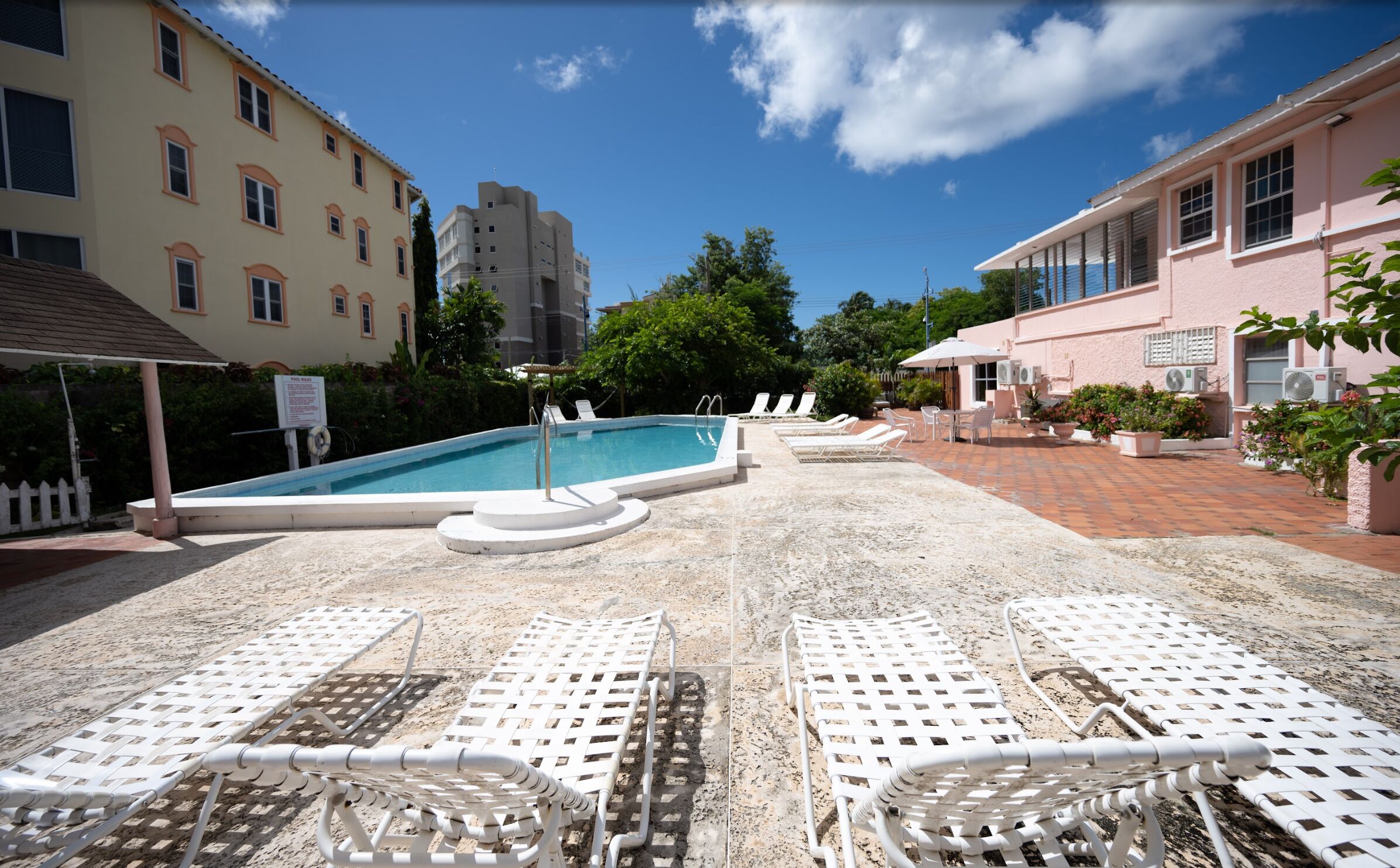 Zona de piscina con sillas reclinables blancas, piscina y edificios al fondo.