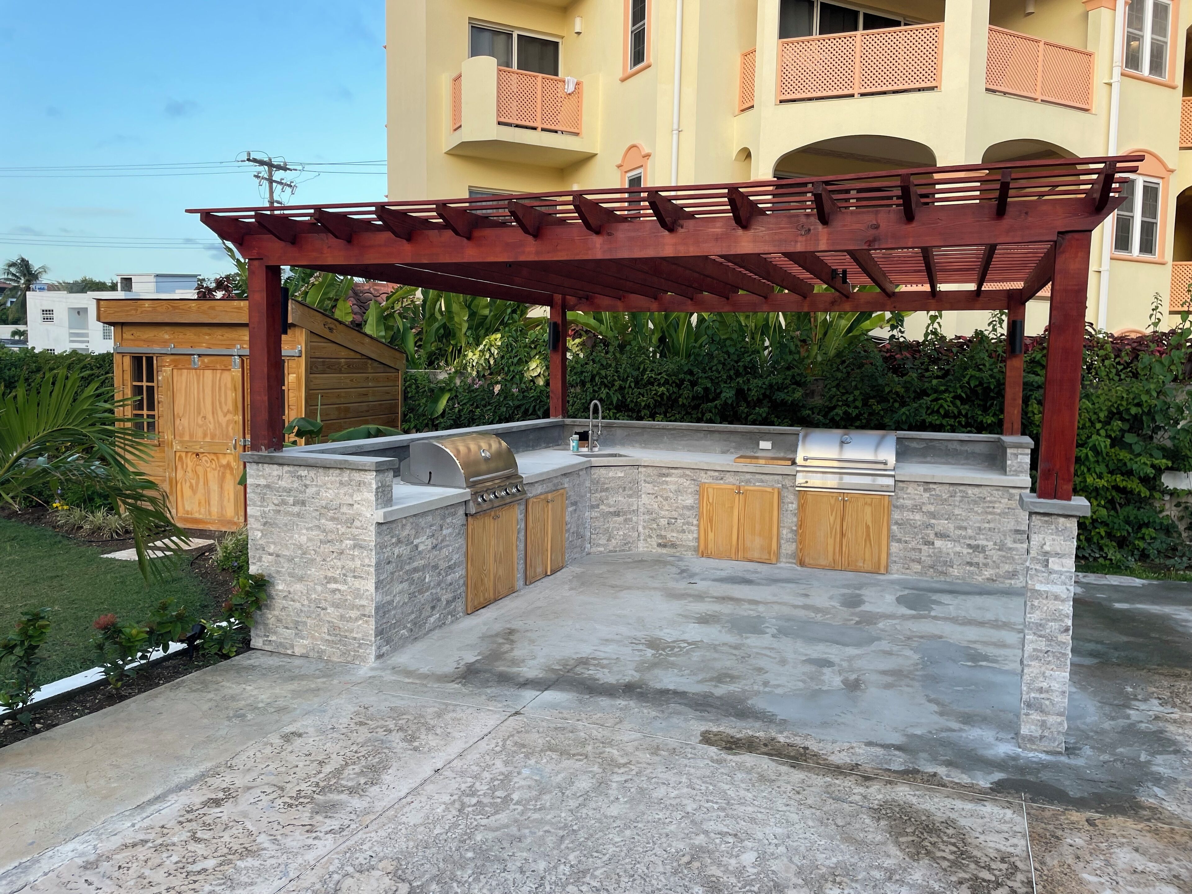 An outdoor kitchen with a stone countertop, wooden cabinets, and a pergola structure.