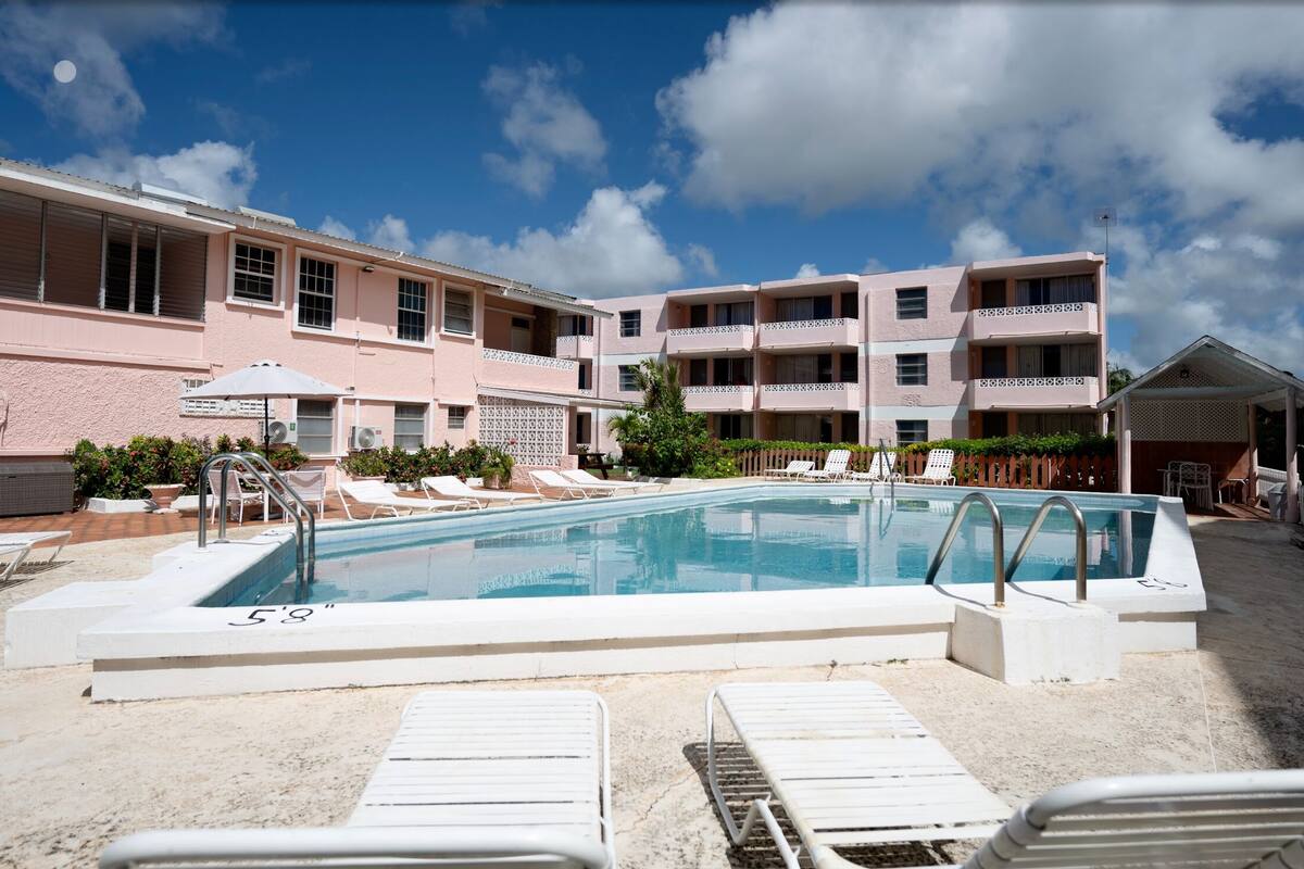 A swimming pool with lounge chairs and a covered area, surrounded by a building with multiple balconies and a clear sky.