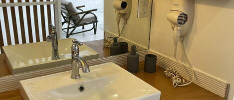 A bathroom with a white sink, wooden vanity, and a mirror. There are two hair dryers mounted on the wall, a towel rack, and a chair visible in the background.
