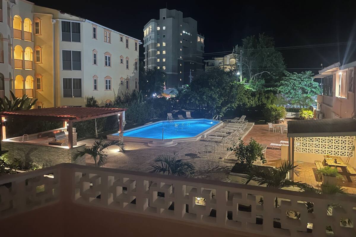 A nighttime view of a residential area with a swimming pool, lounge chairs, and a gazebo.