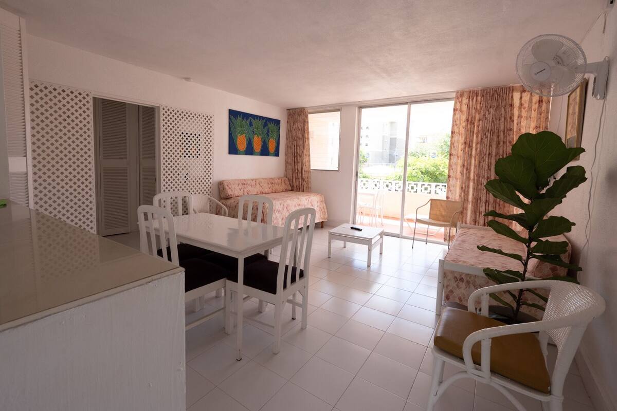 A bright, well-lit room with a dining table, chairs, a sofa, and a balcony with a view.
