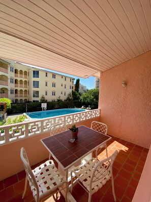 A balcony with a table and chairs, a view of a swimming pool, and a building with balconies in the background. - The Monteray Hotel (St. Lawrence Gap)