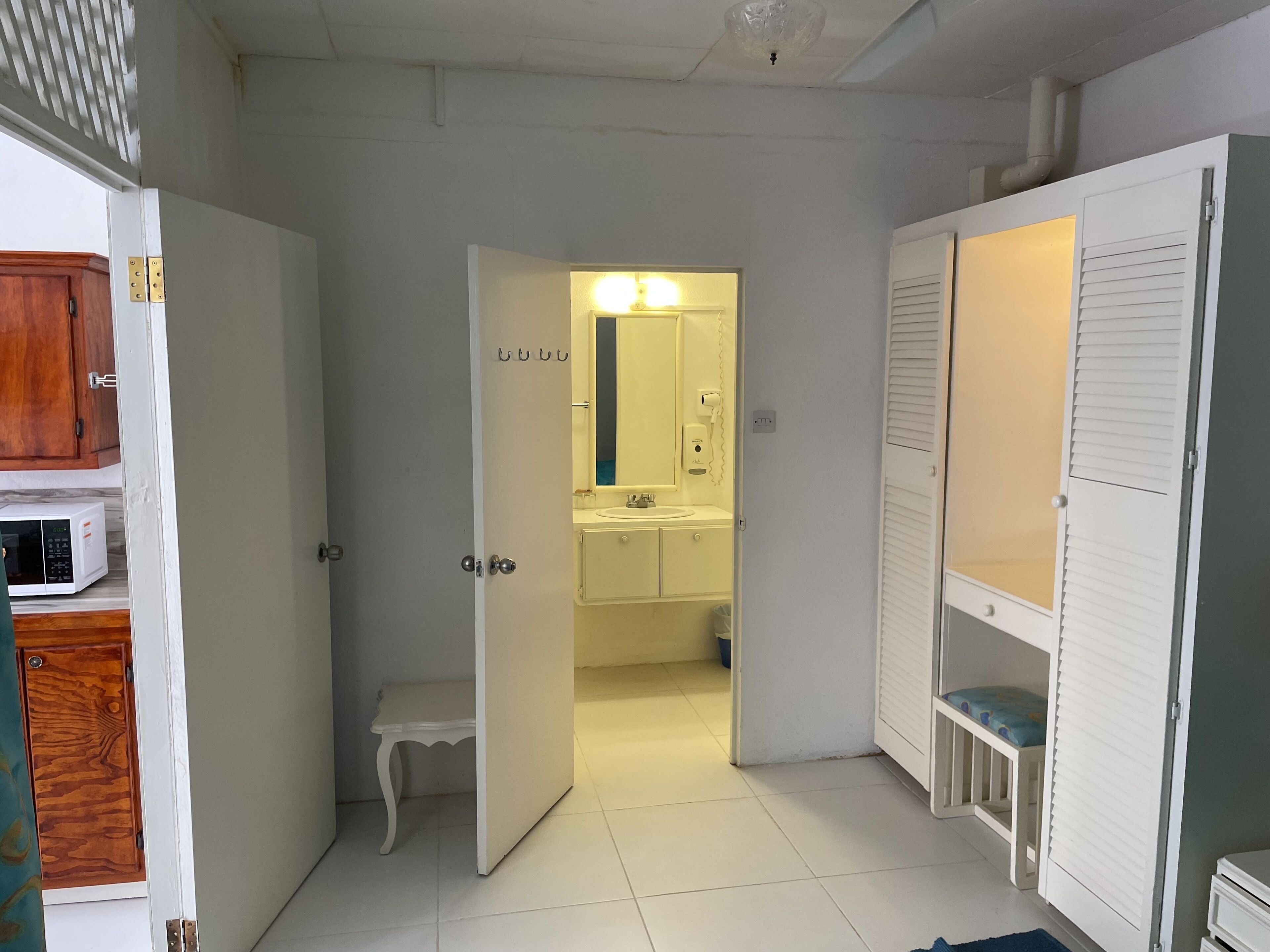 A room with a white door leading to a bathroom with a sink, mirror, and toilet. There is a small white bench and a white cabinet with a microwave on the left.
