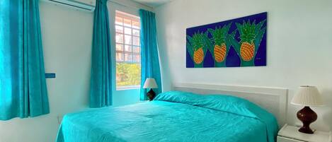 Ein Zimmer mit einem Bett, einem Nachttisch, einer Lampe, einem Bild von Ananas und einem Fenster mit Vorhängen.