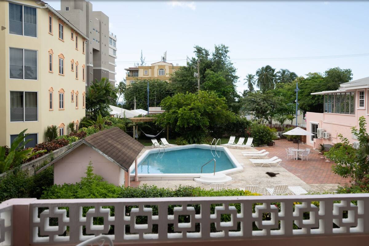 A pool area with lounge chairs and a small table, surrounded by greenery and buildings.