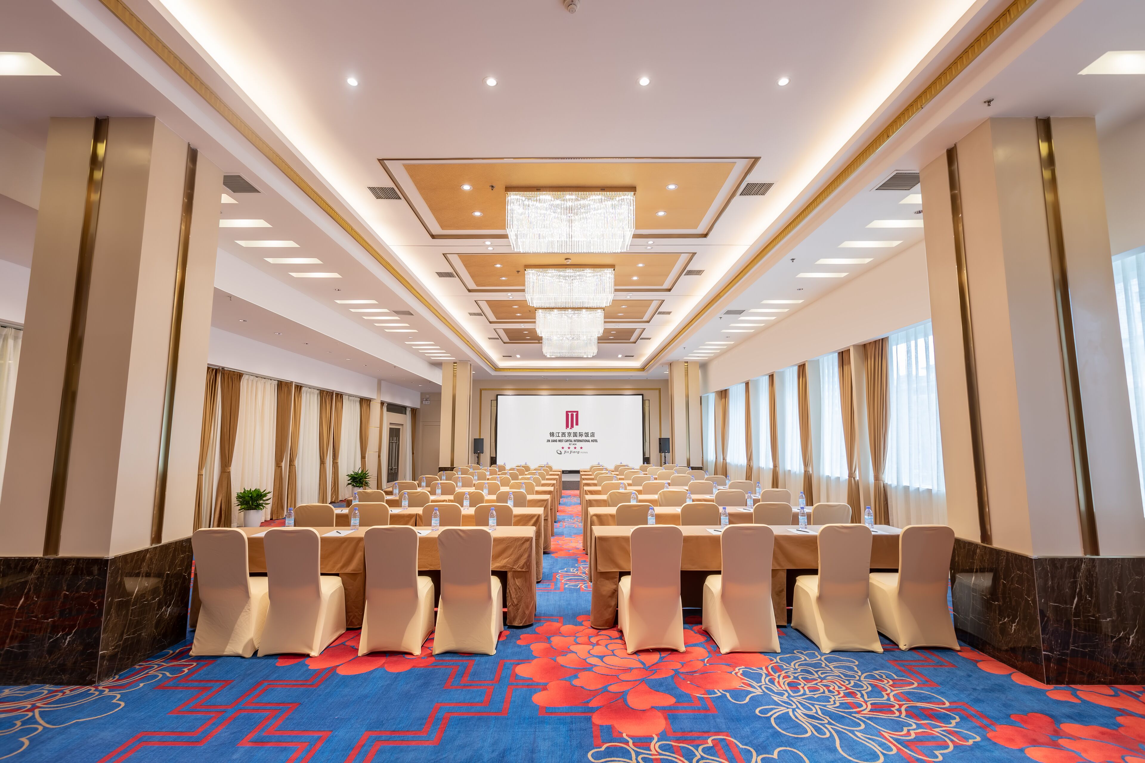 Une salle de conférence avec des rangées de tables et de chaises, un grand écran à l’avant et un tapis à motifs.