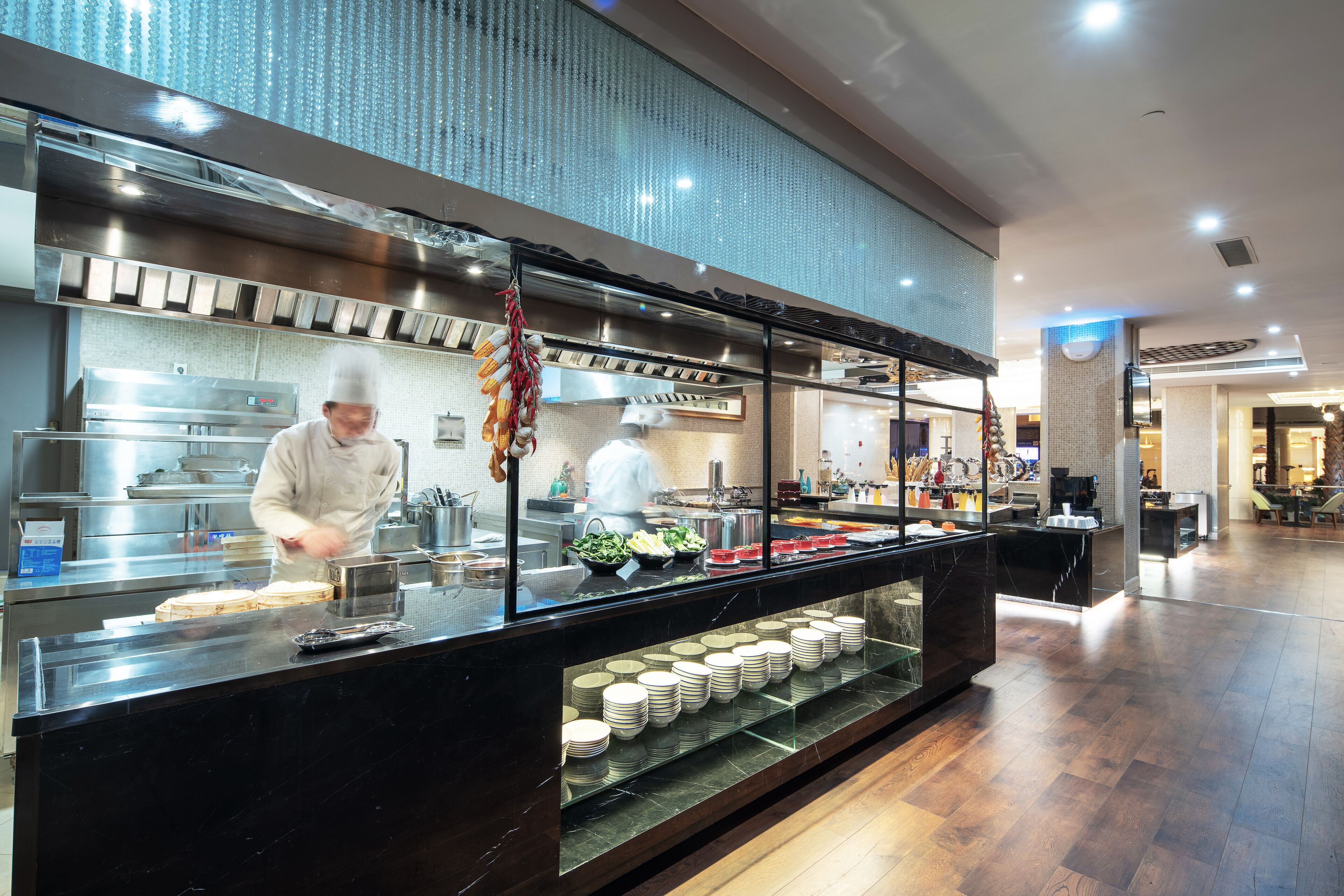 A buffet setup with a chef preparing food, a glass display case with plates, and a modern kitchen in the background.