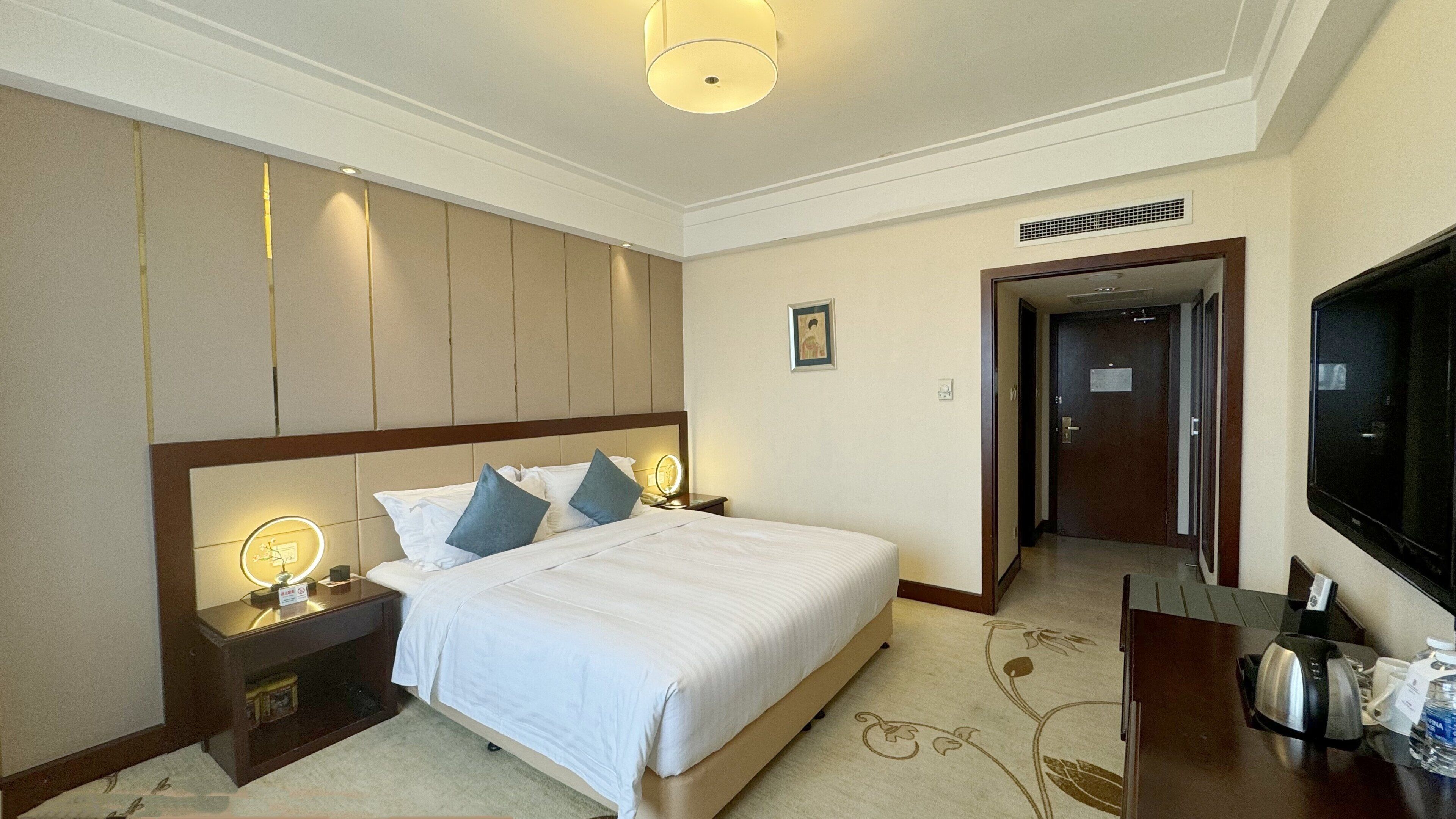 Habitación de hotel con una cama grande, lámparas de noche, un televisor de pantalla plana, una cafetera y una puerta que da a un pasillo.