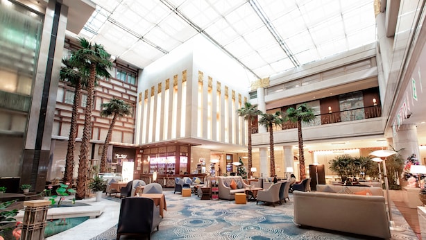 Een ruime lobby met een hoge plafond, palmbomen en een centraal gelegen zithoek met sofa’s en fauteuils.