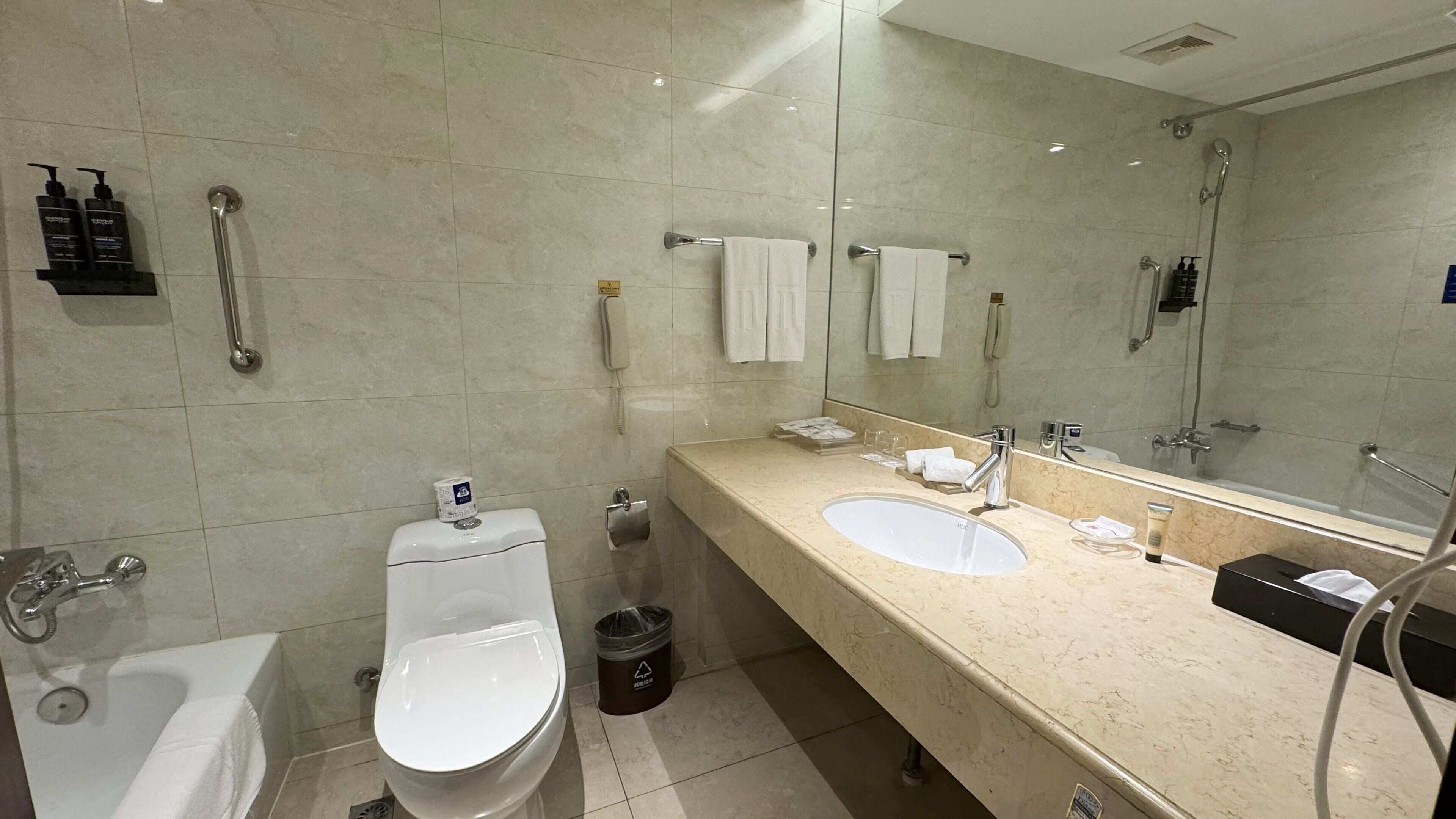 Un baño moderno con inodoro, bidé y lavamanos. Hay barras de apoyo, un espejo y un cubo de basura.