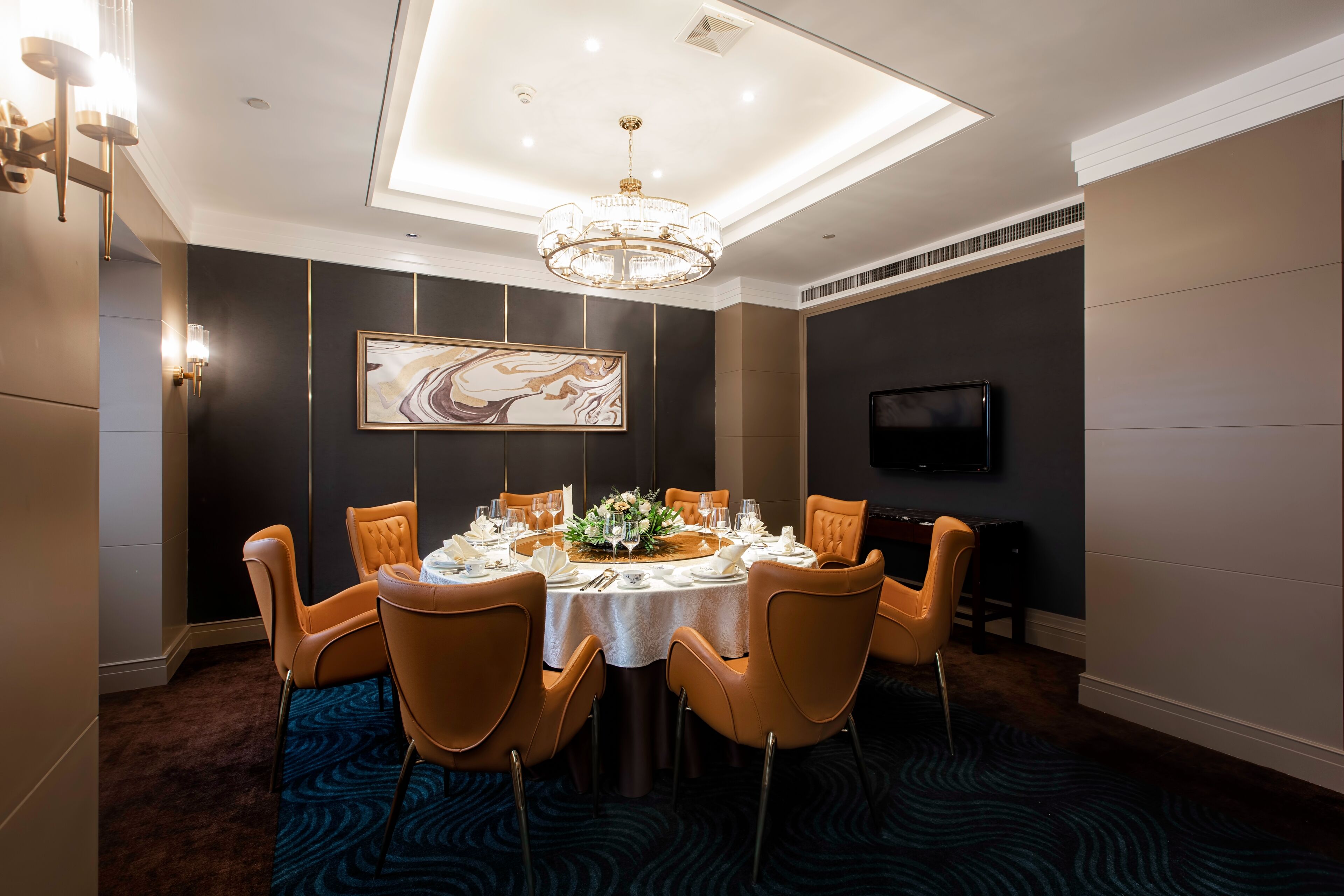 Comedor con mesa redonda para seis personas, sillas naranjas, una lámpara de araña y un cuadro abstracto grande en la pared.
