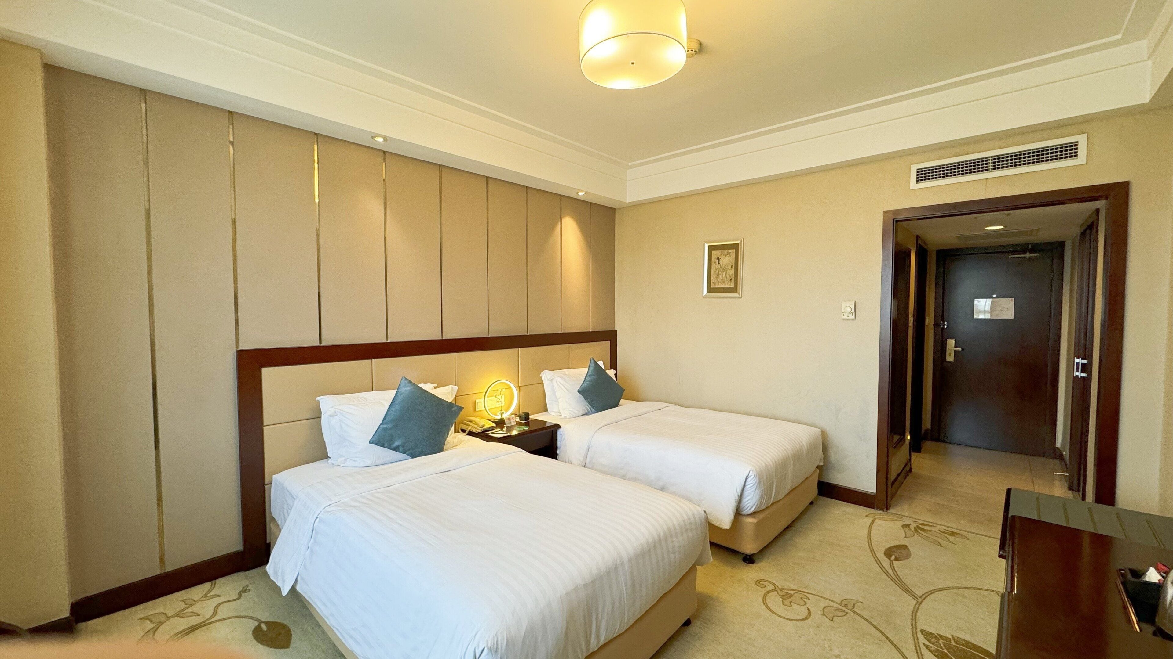 A hotel room with two beds, a nightstand with a lamp, a small table, and a door leading to another room.