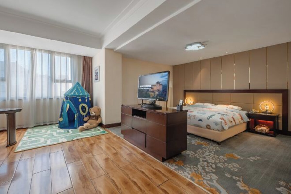 Une chambre d’hôtel avec un plancher en bois, un lit recouvert d’une courtepointe à motifs floraux, un bureau avec une télévision, une petite tente de jeu et une table de chevet avec une lampe.