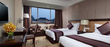 A hotel room with two beds, a desk with a TV, a chair, a lamp, and a view of a building.