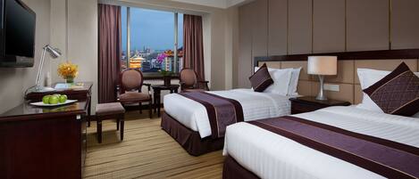 A hotel room with two beds, a desk with a lamp, a chair, a TV, and a view of a cityscape.