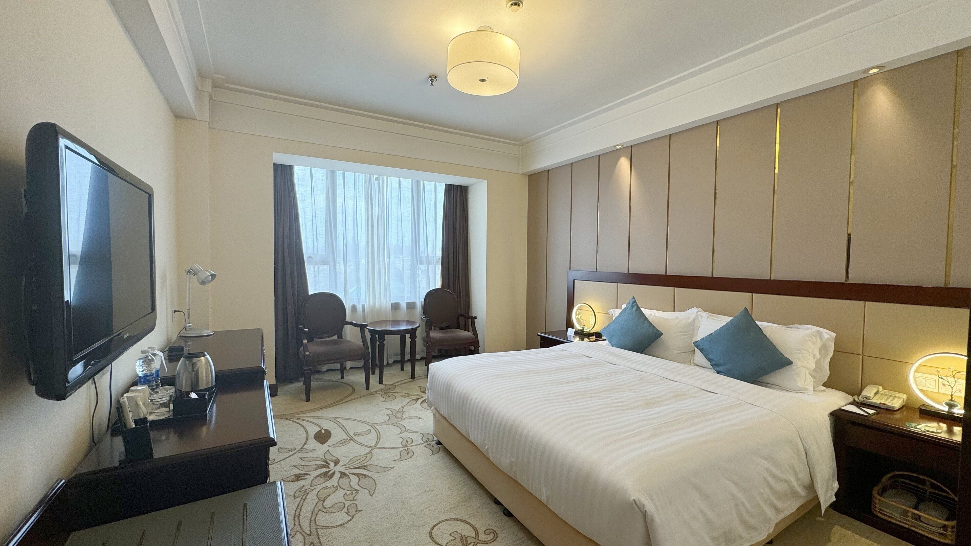 A hotel room with a large bed, a desk with a flat-screen TV, a chair, a small table, and a window with curtains.