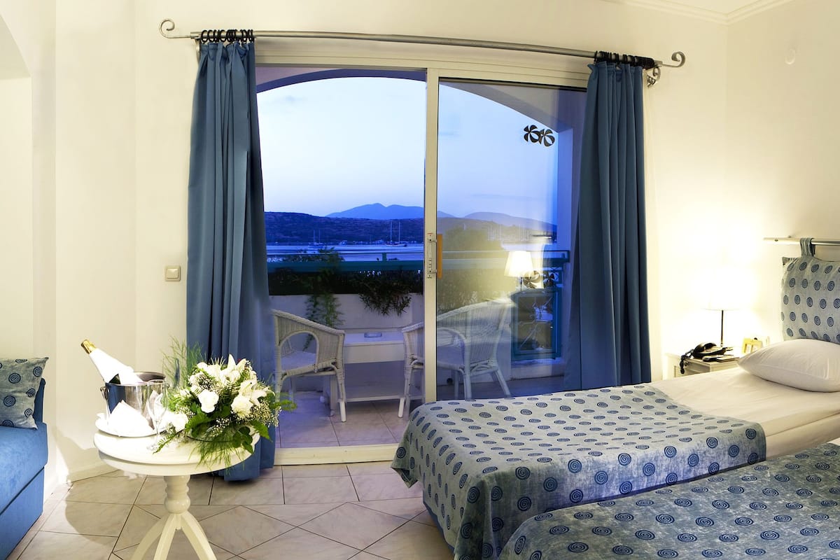 A hotel room with a balcony, two beds with matching blue patterned bedspreads, a small table with a lamp and flowers, and a chair.
