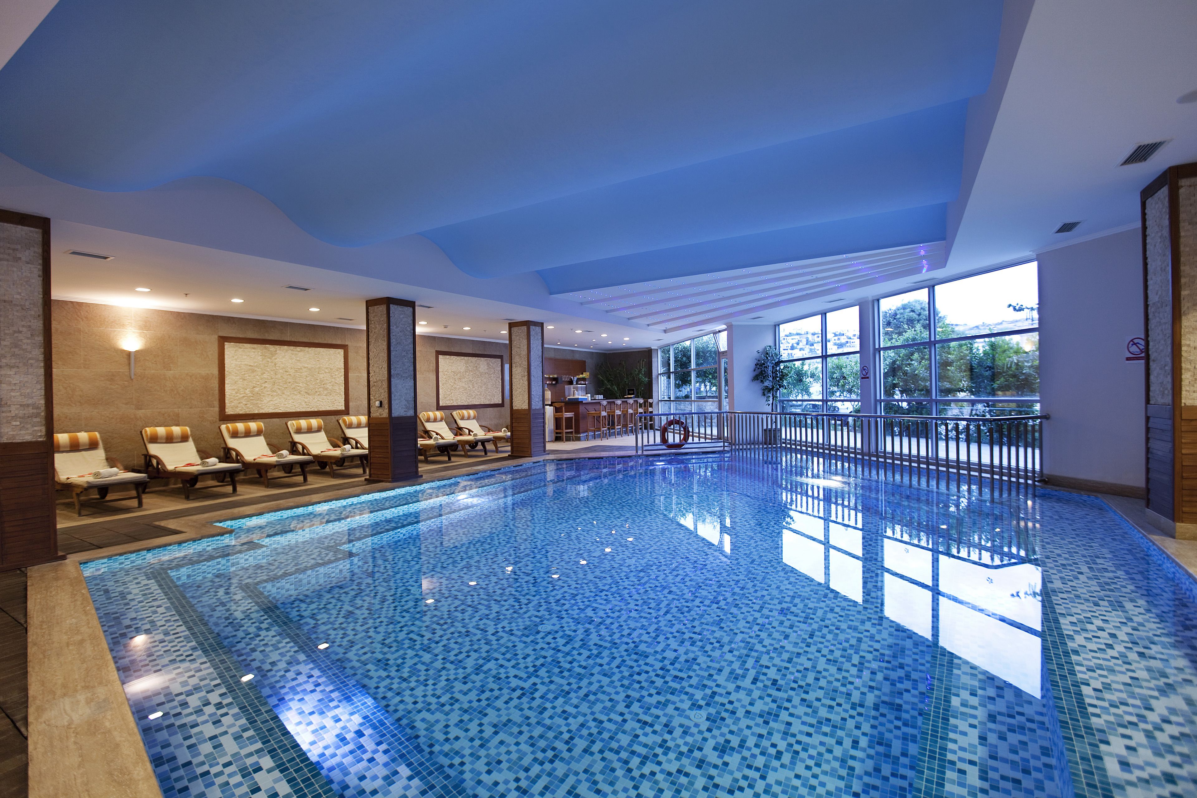 A large indoor swimming pool with tiled flooring, sun loungers along the walls, and large windows providing natural light.