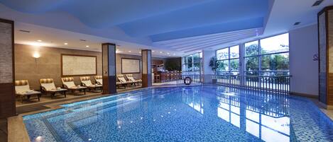 A large indoor swimming pool with tiled flooring, sun loungers along the walls, and large windows providing natural light.