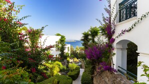 A narrow pathway flanked by lush greenery and flowering plants, leading to a white building with a balcony overlooking a clear blue sky and a distant view of the sea. - Royal Asarlik Beach Hotel & Spa - All Inclusive (Bodrum)