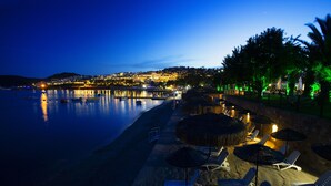 A beachside resort with lounge chairs and umbrellas, a calm sea, and a cityscape in the background. - Royal Asarlik Beach Hotel & Spa - All Inclusive (Bodrum)