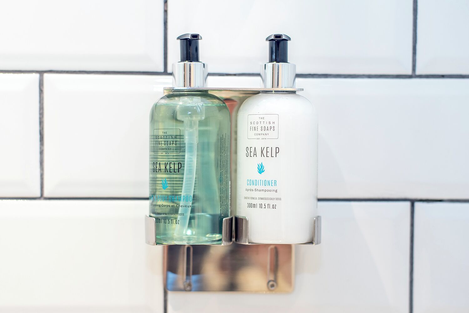 Two bottles of The Scottish Fine Soaps Company products, Sea Kelp Conditioner and Sea Kelp Shampoo, mounted on a white tiled wall.