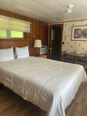 A hotel room with a large bed, a bedside table with a lamp, a small table with a phone, and a door leading to another room. - Starlight Inn (Bennington)