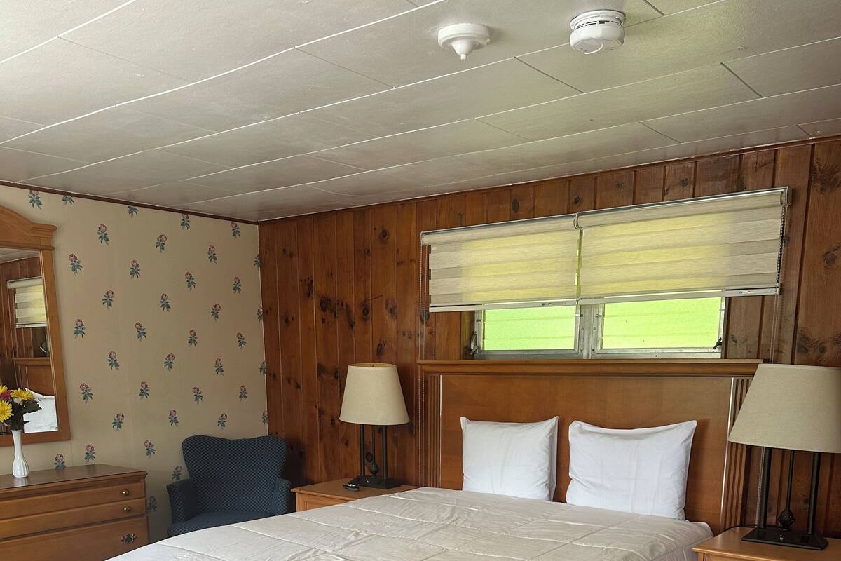 Ett hotellrum med en stor säng, träpanel på väggarna, ett fönster med persienner, ett nattduksbord, en lampa och en byrå.