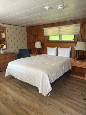 A hotel room with a large bed, wooden paneling, a window with blinds, a nightstand, a lamp, and a dresser.