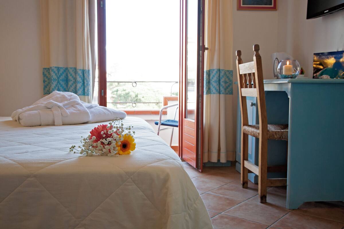 A hotel room with a bed, a desk, a chair, a TV, and a balcony with curtains.