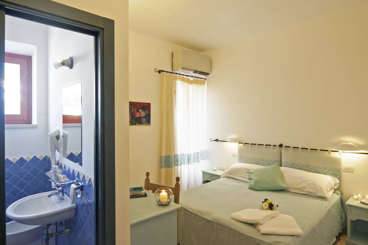 A bedroom with a bed, a nightstand, a bedside table, a window with curtains, and a bathroom with a sink and mirror.
