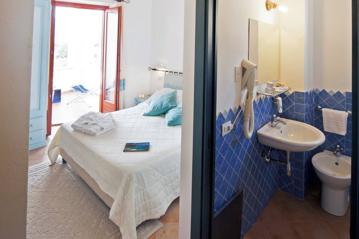 A bedroom with a bed, a bedside table, and a window. The room has a white bedspread and pillows. There is a bathroom with a sink, a mirror, and a bidet.