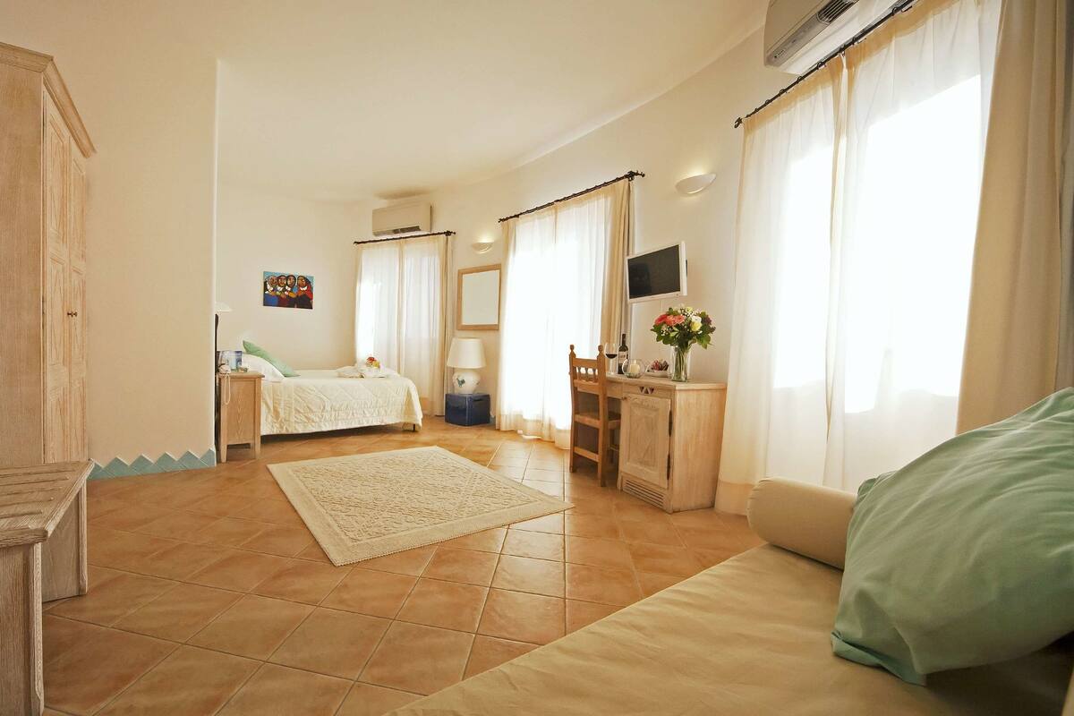 A well-lit, spacious room with a bed, a desk, a chair, and a sofa. There is a small table with a lamp and a vase of flowers.