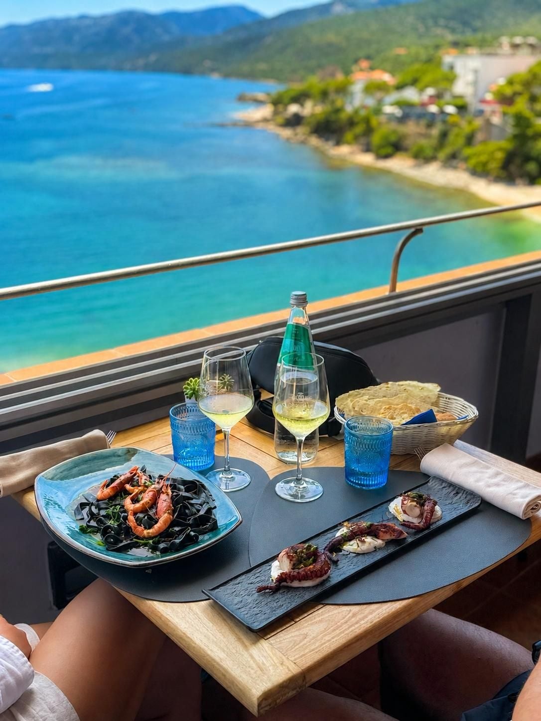 A dining table with seafood dishes, glasses, and a bottle, overlooking a coastal view.