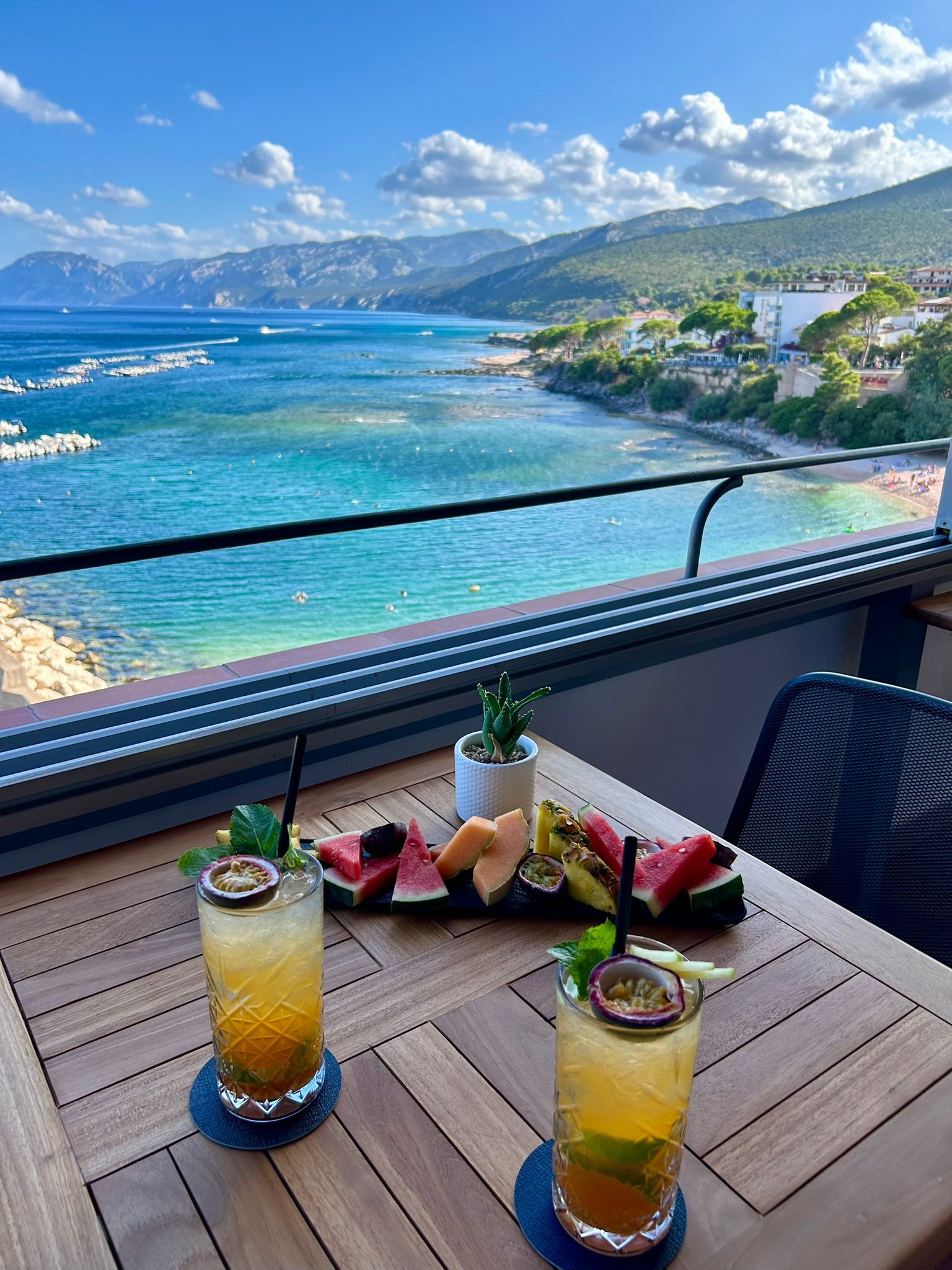 Une table avec deux boissons et une assiette de fruits sur un balcon donnant sur la côte.