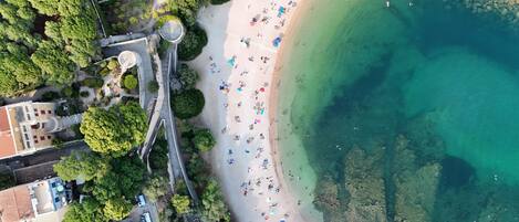 Une vue aérienne d’une plage où l’on aperçoit des personnes, une zone d’eau claire et une végétation luxuriante environnante.